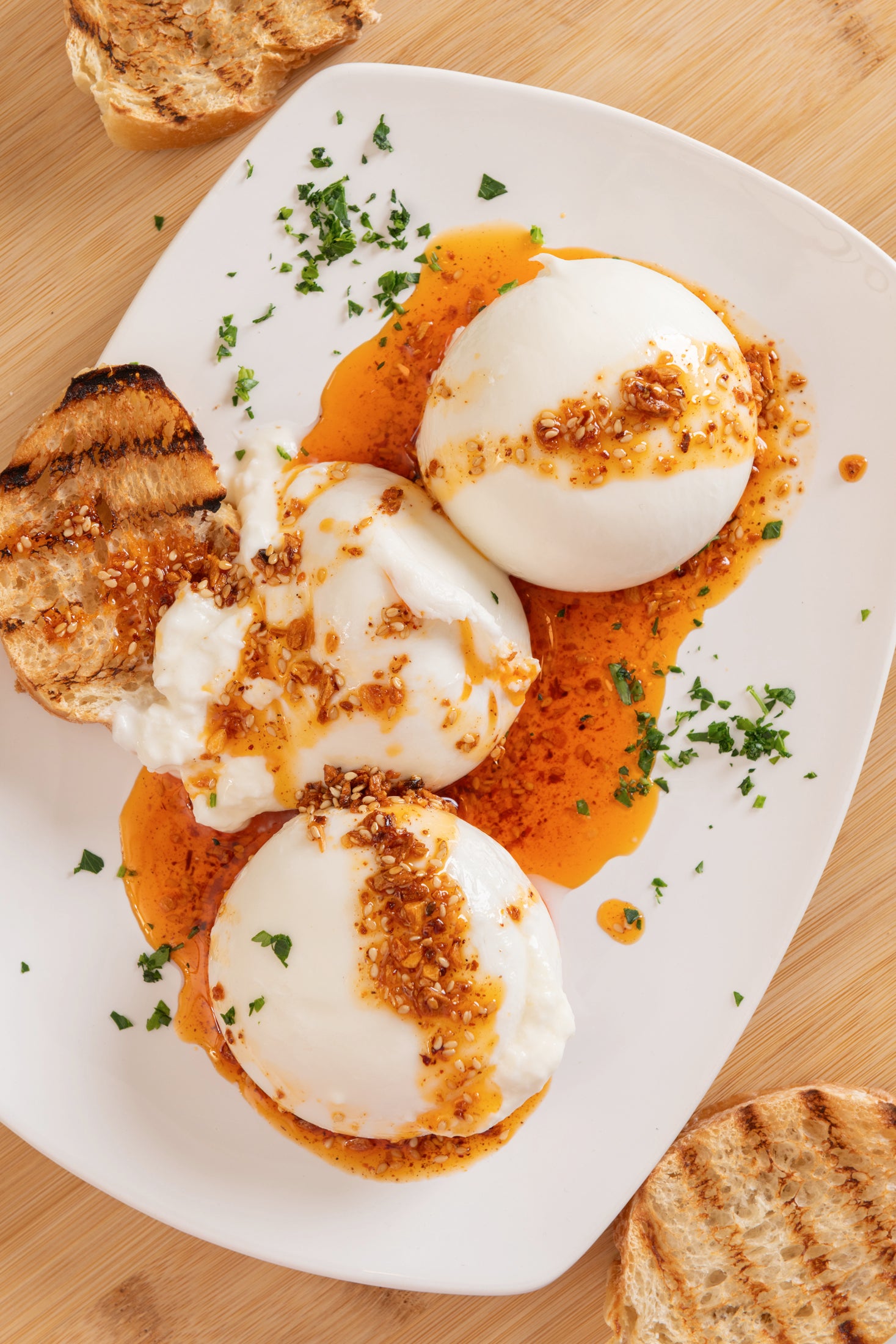 Ruby Crunch chili crisp pooled around and drizzled on top 3 balls of burruta cheese with grilled toast garnished with minced fresh parsley