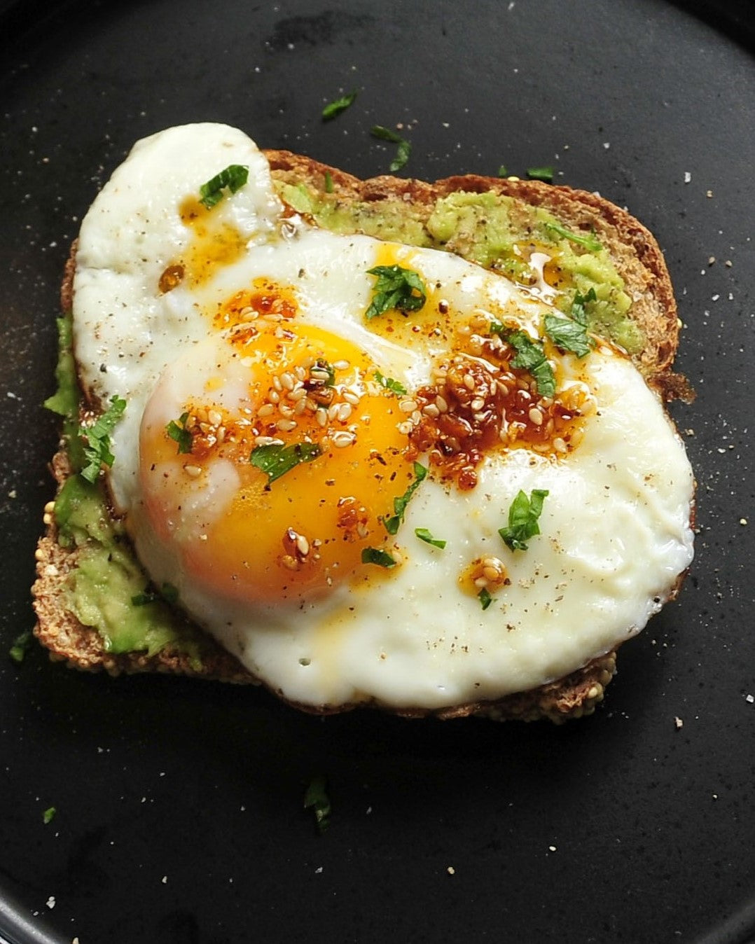 Ruby Crunch chili crisp spooned onto a sunny side up egg set on a piece of grainy toast spread with smashed avocado