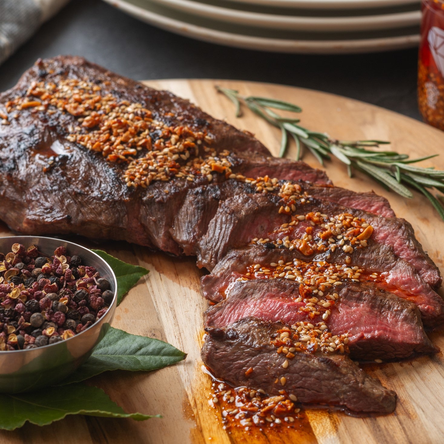 Ruby Crunch chili crisp on a slices of flank steak on a wooden cutting board accompanied by rosemary, peppercorns and bay leaves