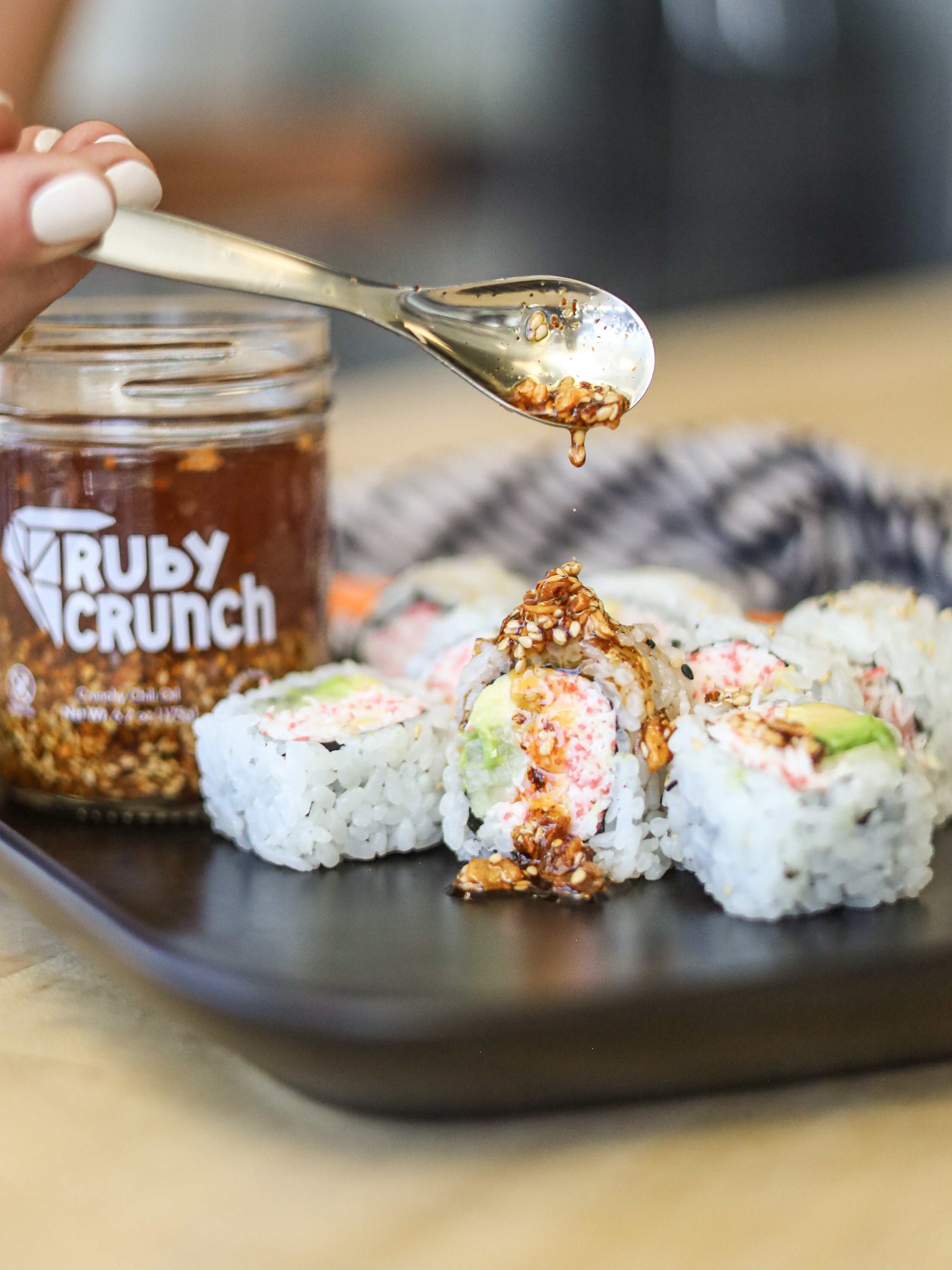 Ruby Crunch chili crisp drizzling onto a sushi role on black plate with a jar of Ruby Crunch chili crisp in the background