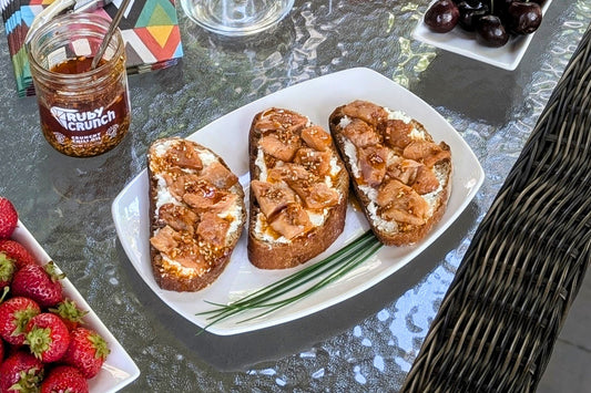 Appetizer plate with fresh bakery bread and soft cheese, topped with tinned fish and a drizzle of Ruby Crunch chili crisp. Strawberries and cherries are visible as summer snacks.
