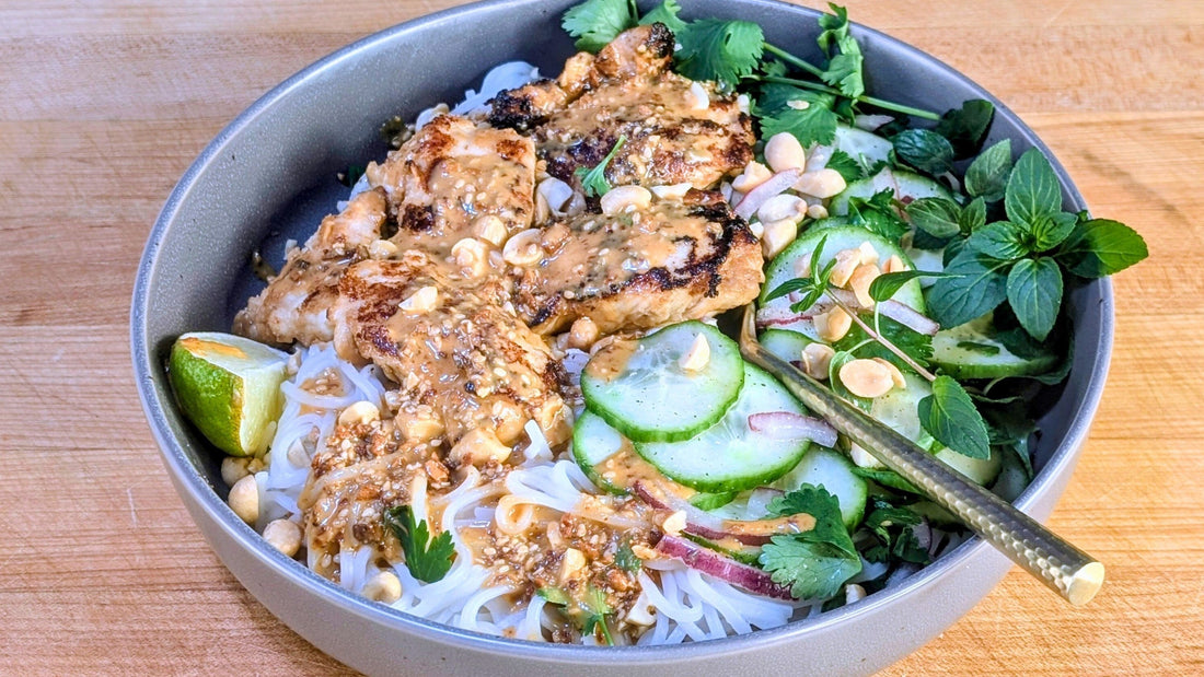 A bowl with rice noodles, chicken, unami peanut sauce, cucumbers and herbs with crushed peanuts.