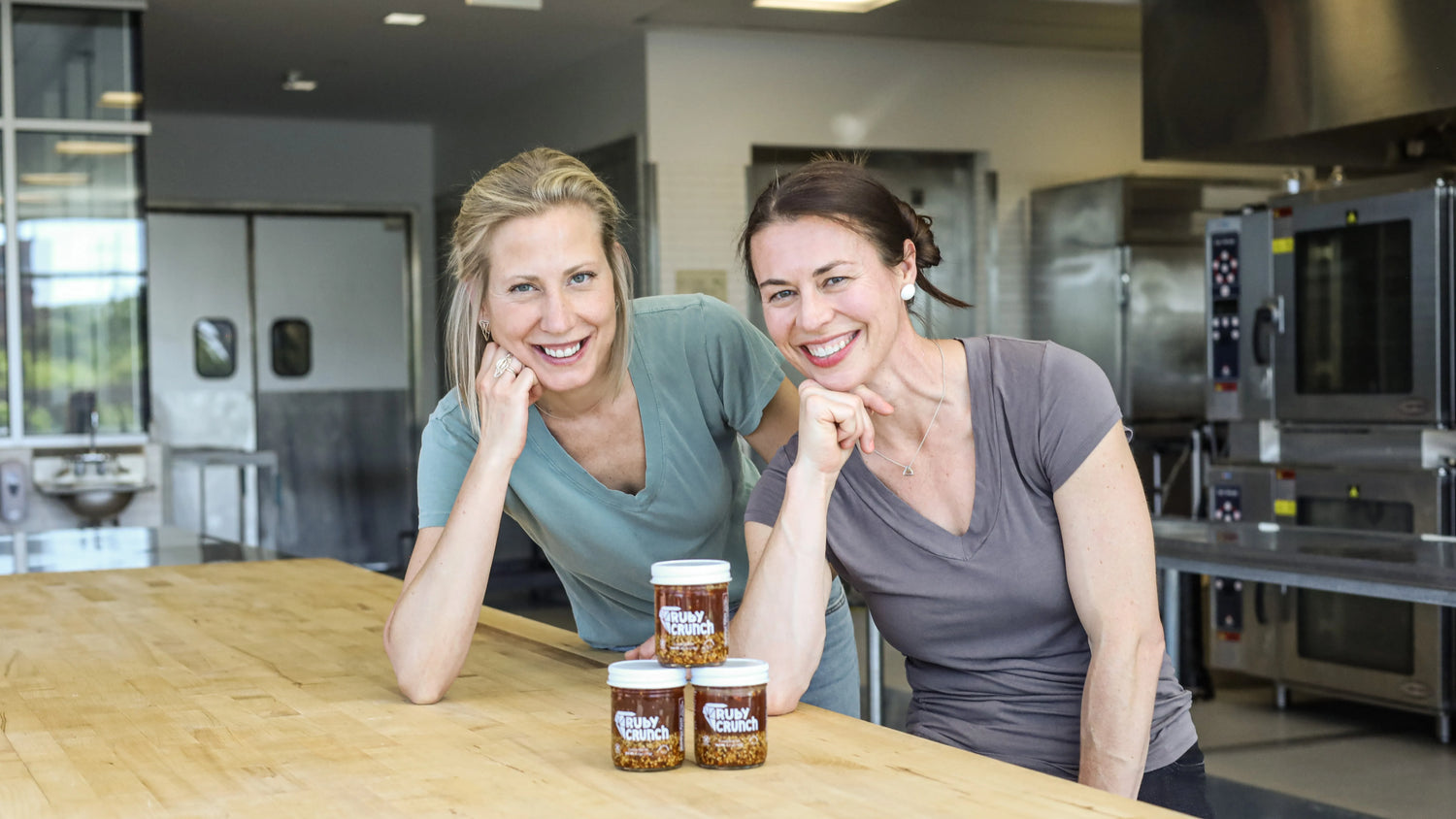 Two women, the founders and makers of Ruby Crunch chili crisp, in a commercial kitchen leaning over a count with their product Ruby Crunch chili crisp