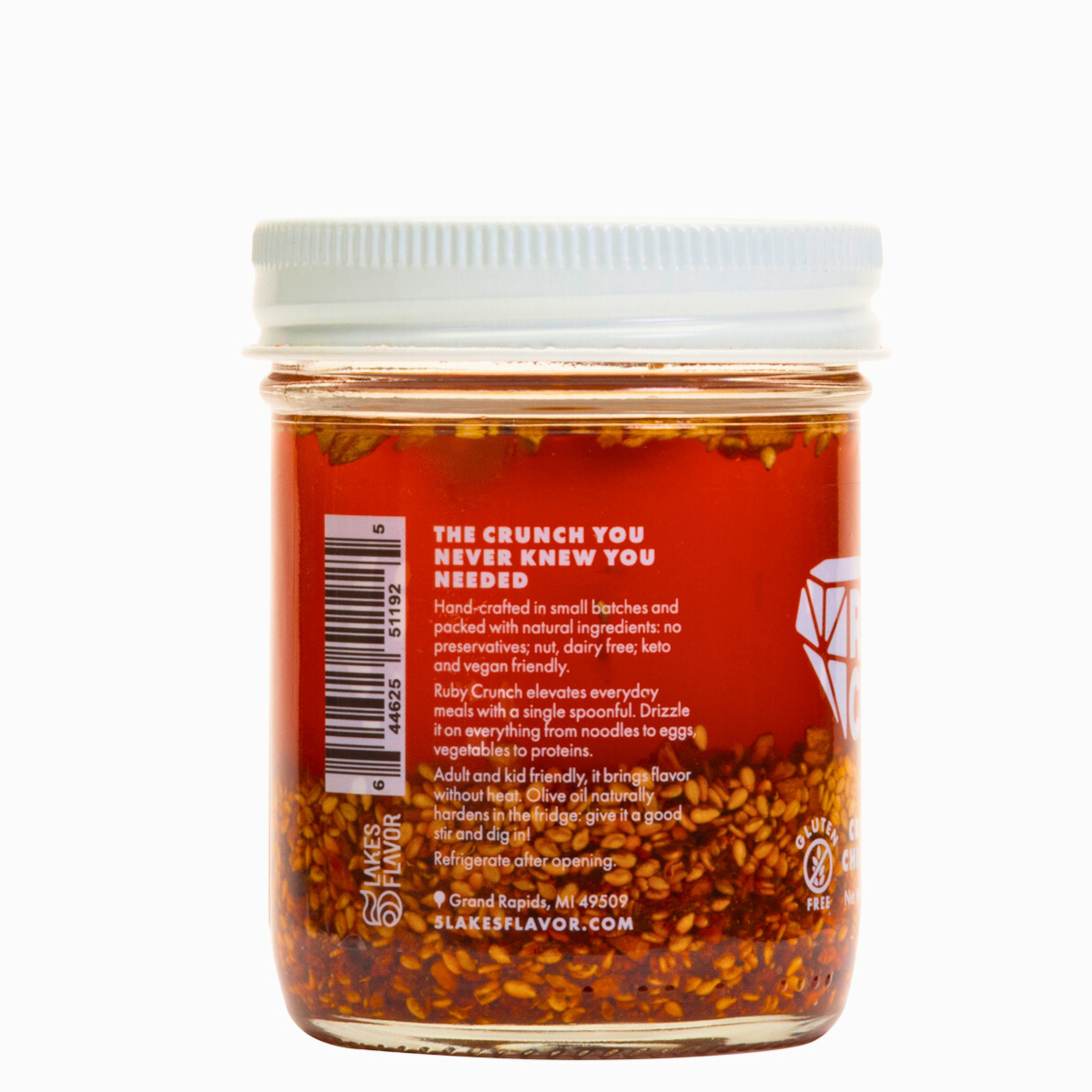 Side view of Ruby Crunch olive oil chili crisp condiment jar, with the product description, showing the ruby hued oil & crispy bits inside.