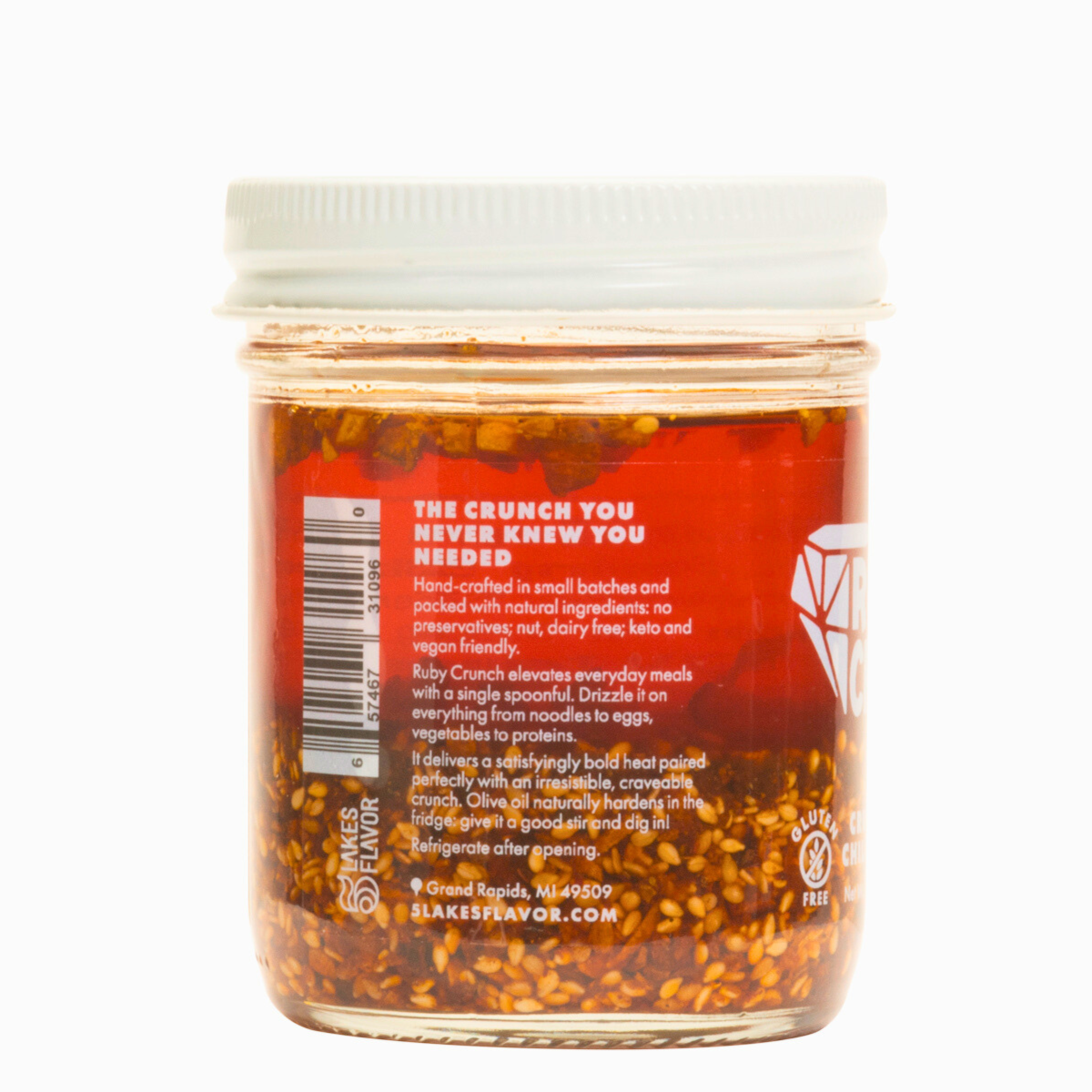 Side view of Spicy Ruby Crunch olive oil chili crisp condiment jar, with the product description, showing the ruby hued oil & crispy bits inside.